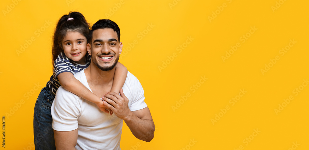 Fototapeta premium Portrait Of Happy Young Middle Eastern Dad And Cute Little Daughter Posing Together Over Yellow Background, Female Kid Embracing Daddy From The Back And Smiling At Camera, Panorama, Copy Space