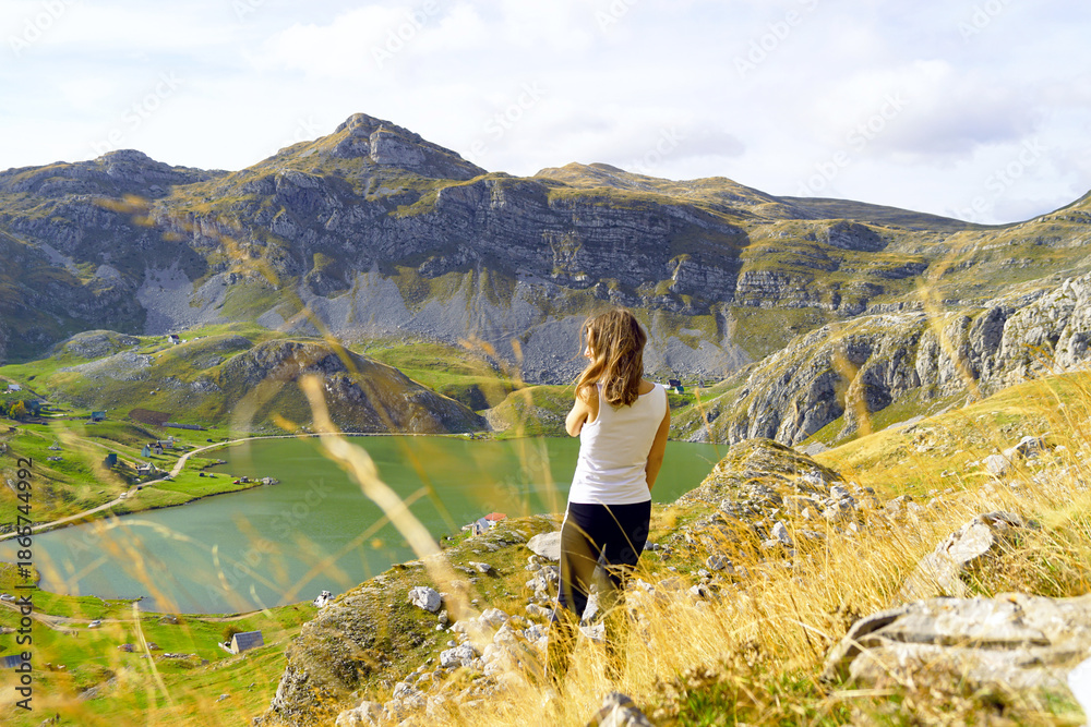 Fototapeta premium Woman standing on slope near alpine lake in Moraca mountains, Montenegro. Represents reflective travel mood, solo hiking lifestyle, quiet observation of nature, slow tourism trend, and personal space.