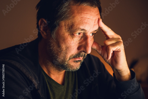 Concerned man looking at computer screen and thinking in dark room illuminated by warm low key light.