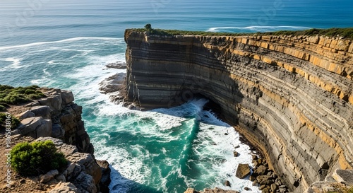 Dramatic rugged coastal cliffs meet turbulent turquoise ocean waves under a clear blue sky.