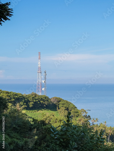 Cell Tower on Lush Green Hilltop Overlooking Blue Ocean Under Bright Sky, Offering Cellular Connectivity in Coastal Regions for Modern Communication