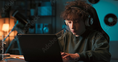 Quadro em tela Smiling young college-aged boy has wireless headphones listens to music and works at computer, writes paper for school at desk at home