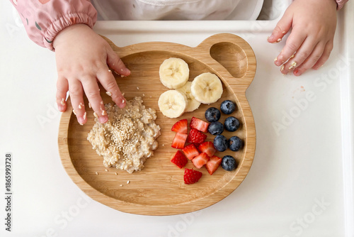 Caucasian baby learning self feeding with oatmeal, banana and berries on wooden plate. Baby led weaning concept, healthy toddler nutrition and safe home mealtime routine.