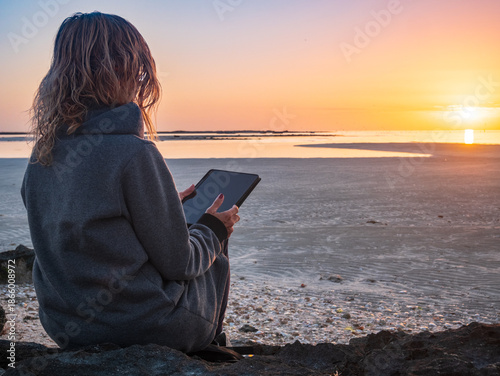 Woman using tablet on quiet beach in winter at sunset, back view, calm seascape in Djerba Tunisia, remote work, solitude, travel lifestyle