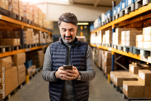 Middle-aged warehouse manager using smartphone in storage. Professional businessman in distribution center. E-commerce, supply chain and technology concept. Logistics worker.