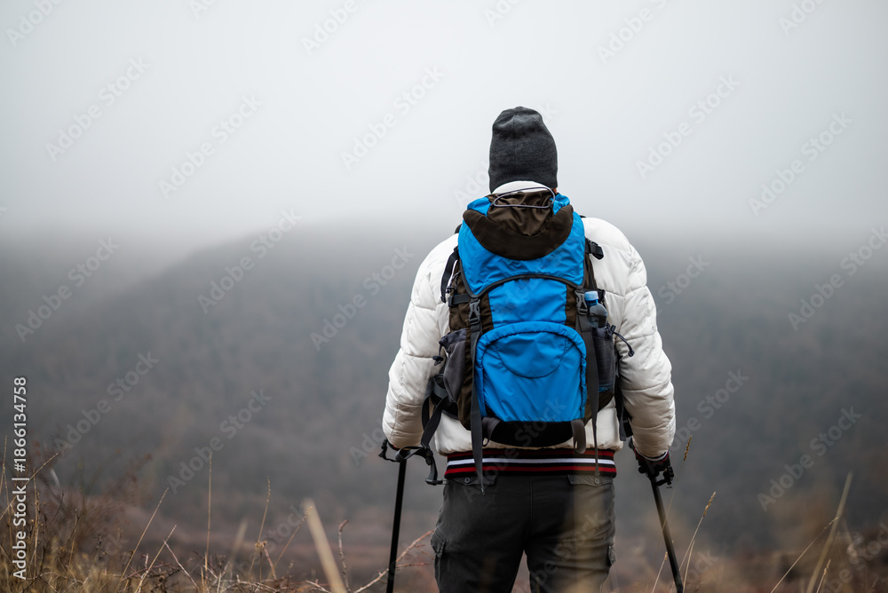 Naklejka premium Rear view of hiker in warm clothing with backpack and hiking poles enjoying the atmospheric winter fog and the peacefulness of nature while on his hiking journey.
