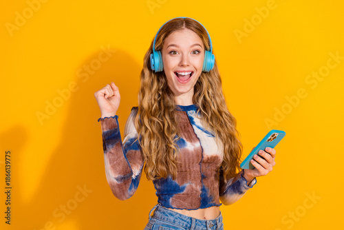 Young woman with blue headphones smiles while holding a phone against a bright yellow background showcasing casual fashion and music lifestyle