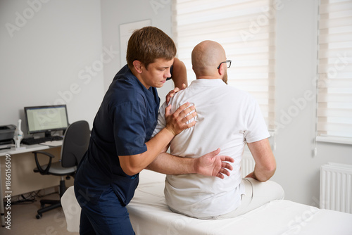 Chiropractor adjusting male patient’s shoulder in clinic. Physiotherapist performing manual therapy to relieve pain, improve mobility, support rehabilitation after injury or musculoskeletal disorder.