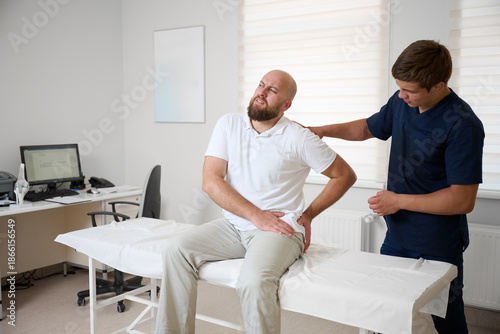 Man suffering from hip pain during medical consultation in physiotherapy clinic. Patient holding leg in discomfort while doctor examines his hip. Concept of joint injury, rehabilitation, orthopedic tr