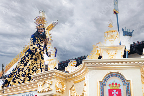 Konstfotografi Ornate statue of Jesus with golden crown and embroidered robe, carrying the cross on a float during a Semana Santa procession under cloudy skies