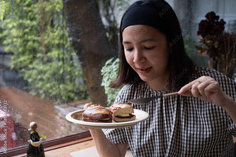 Φωτογραφία Asian woman enjoying pretzel sandwich in cozy cafe by rainy window, holding plate and knife with natural light