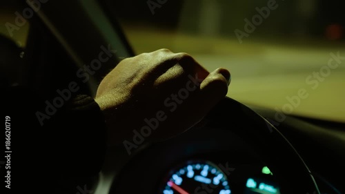 Hand resting on steering wheel while unrecognizable man driving on highway at night. Male cruising road, gripping wheel under street lighting. Nighttime driving along expressway, steering vehicle