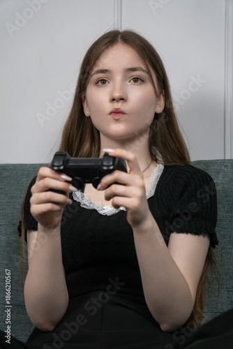 Teenager sitting comfortably on a sofa at home, fully immersed in playing video games with a wireless game controller, enjoying leisure time and relaxation. Vertical photo