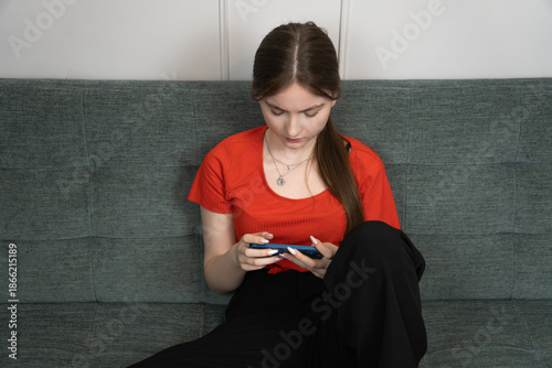 Sad teenage girl using smartphone sitting on sofa at home, experiencing cyberbullying, online harassment, or social media addiction, feeling lonely, anxious, and depressed