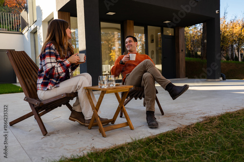 Happy couple sitting outdoors on a modern patio next to their house, drinking coffee and having a pleasant conversation, embracing a relaxed autumn lifestyle