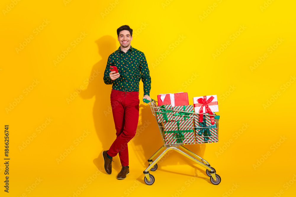 Fototapeta premium Young man with a shopping cart full of Christmas gifts smiles against a bright yellow background