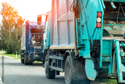 Fotografía Side and rear view of two turquoise and blue garbage trucks on asphalt road, gre