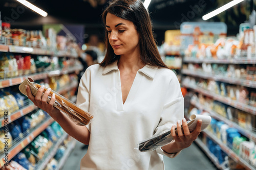Tablou pe pânză Woman in white shirt is comparing two products in a grocery store aisle, surroun