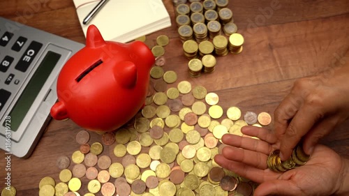Top view of a person managing finances, counting euro coins on a wooden desk with a calculator, notebook, and a red piggy bank nearby