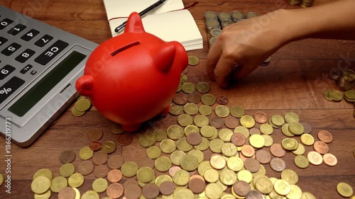 Overhead view of a woman counting and stacking euro coins on a wooden table. A red piggy bank, calculator, and notebook are on the desk