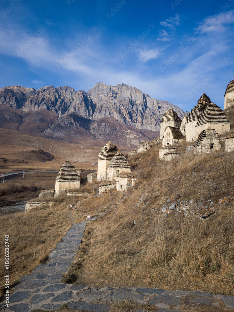 Fototapeta premium Ancient stone structures on a hillside, surrounded by rugged mountains and clear blue sky, showcasing historical architecture and natural beauty in a serene landscape