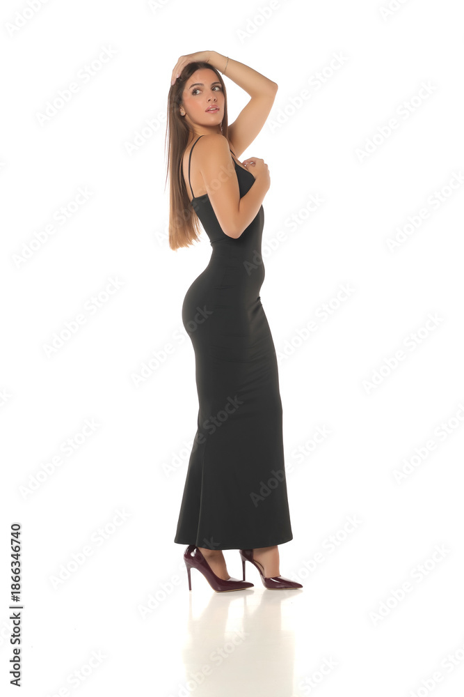 Naklejka premium Studio shot of a woman with dark hair wearing a long black dress and high heels, posing in side view with hand on head, full body, on a white background