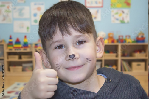 Close-up portrait of a little boy with cat face painting posing in a playroom with toys on the background.