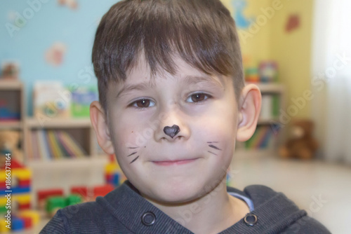 Close-up portrait of a little boy with cat face painting posing in a playroom with toys on the background.
