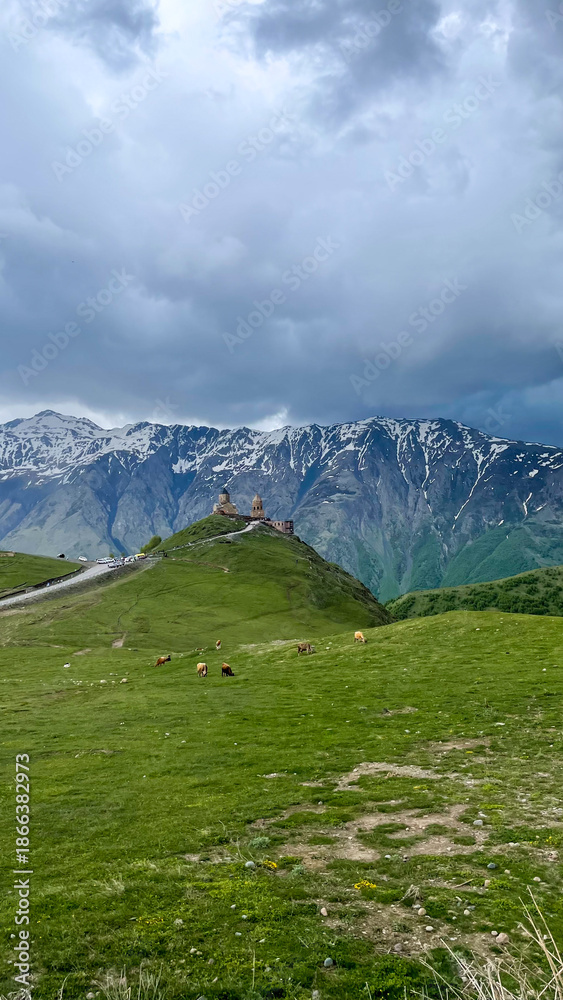 Fototapeta premium Majestic snow-capped peaks cradle ancient chapel under brooding clouds, echoing Caucasus mythology and serene Mountain Day introspection