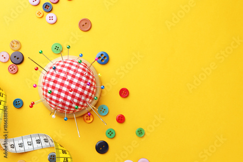 Принти на полотні Checkered pincushion with pins, needles, measuring tape and buttons on yellow background, flat lay