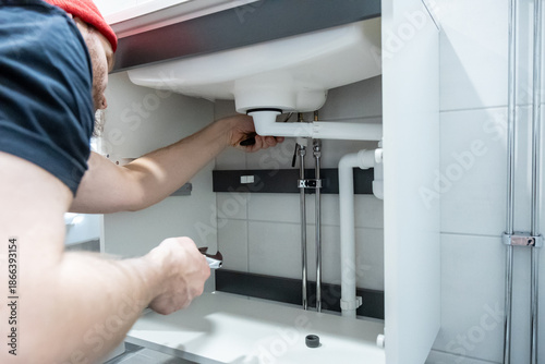 Handyman installing a new faucet and plumbing lines under a bathroom sink in a modern bathroom with tiled floor. Home renovation scene showing repair, maintenance, and proper installation work