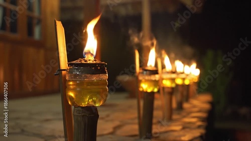 Row of glowing homemade oil lamps made from plastic bottles and bamboo illuminating a path at night with a wooden building in the background