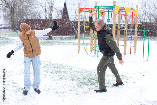 Two young boys play in the snow at a playground. One boy wears a brown vest and the other a black jacket. Snow falls gently around them.