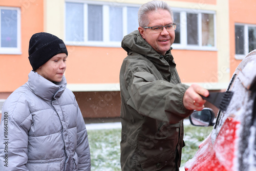 A senior Caucasian man and a young boy are cleaning snow off a car in a snowy environment. The man wears a green jacket and glasses, while the boy is in a gray coat and black beanie.