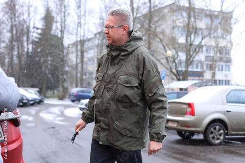A middle-aged Caucasian man with short gray hair wearing a green jacket stands outside near parked cars in a residential area on a cloudy day.