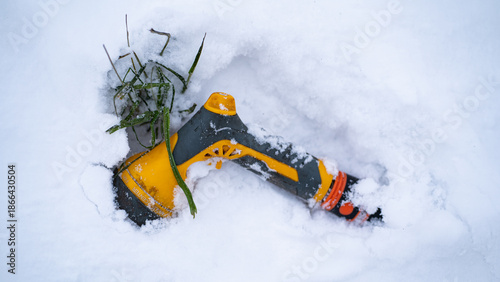Garden hose nozzle partially buried in fresh snow with frozen grass nearby. Concept of winter season, outdoor equipment left outside, frost damage risk and cold weather conditions.