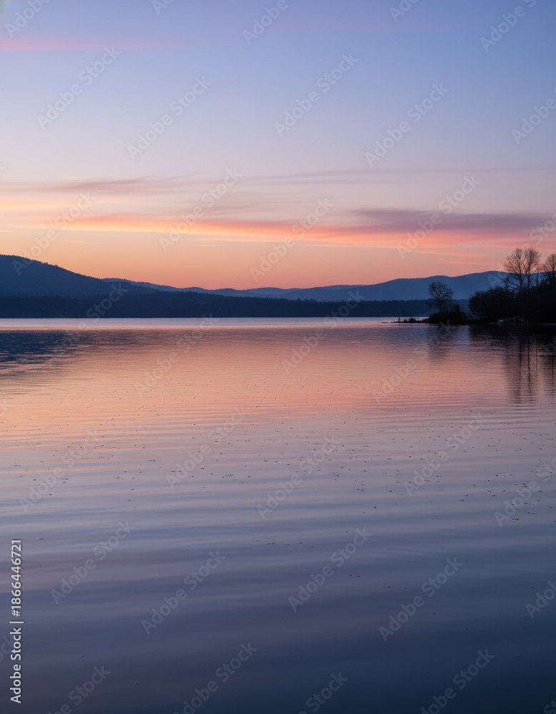 Fototapeta premium Serene lakeside landscape reflecting a vibrant sunset over distant mountains during dusk