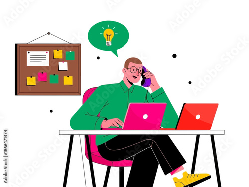 A man sitting at a desk working on two laptops while on a phone call in a modern office setting with a bulletin board around him