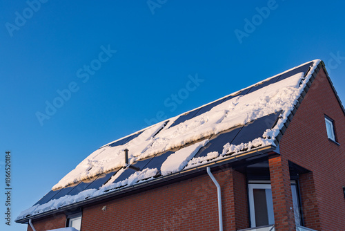 Residential brick house roof with solar panels partially covered in snow against a clear blue sky, reducing energy efficiency.
