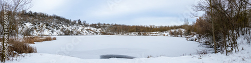 Frozen quarry surrounded by leafless trees and snowy hills under a cloudy winter sky, creating a calm and expansive cold-season landscape
