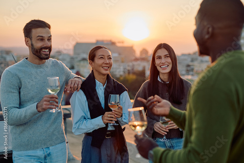 Schilderij op canvas Diverse group of smiling friends enjoying wine and chatting on a city rooftop wi