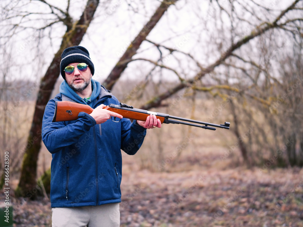 Fototapeta premium A man in a green jacket and hat is holding a rifle. He is smiling and looks confident handling the weapon. Hunter holding light small caliber gun. Grey winter snowless background.