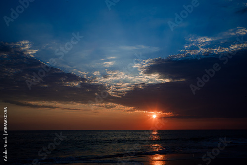 Seascape during sunset featuring dark clouds and golden sunlight reflecting on the wet sand of the shore.