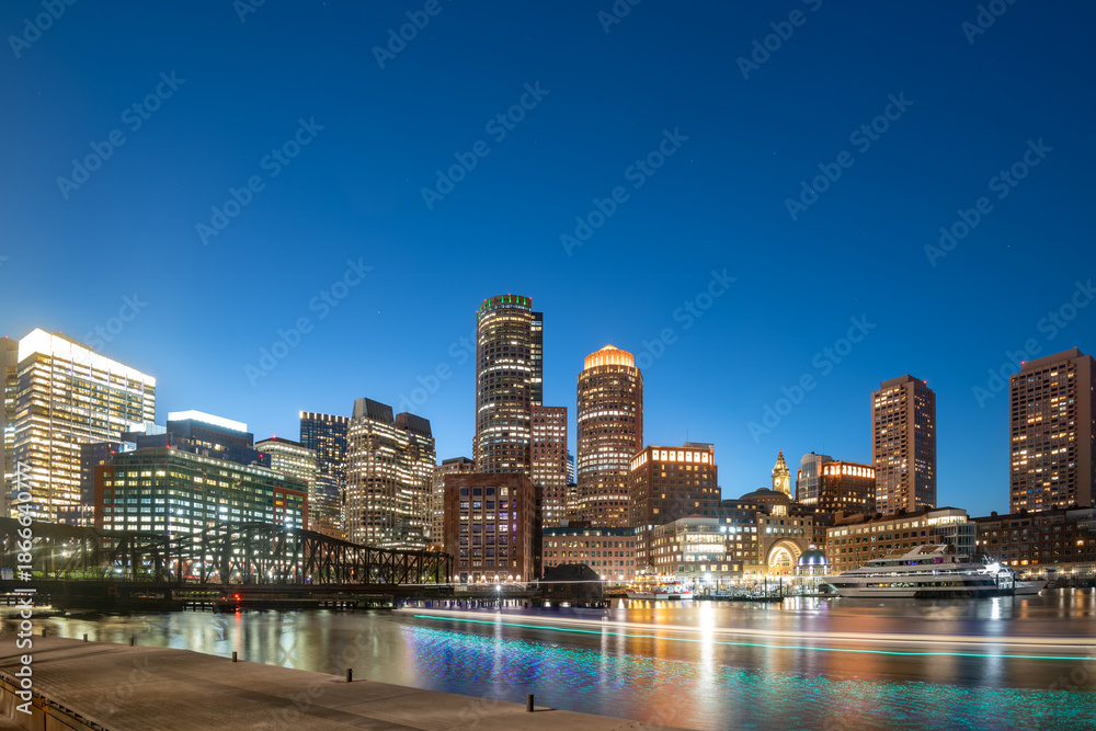 Obraz premium A stunning view of the Downtown Boston skyline at night featuring illuminated skyscrapers and golden reflections on the water.