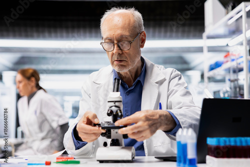 Senior lab manager checks product surface for tiny defects using microscope. Elderly research facility professional ensures manufacturing standards, examines small parts with optical device