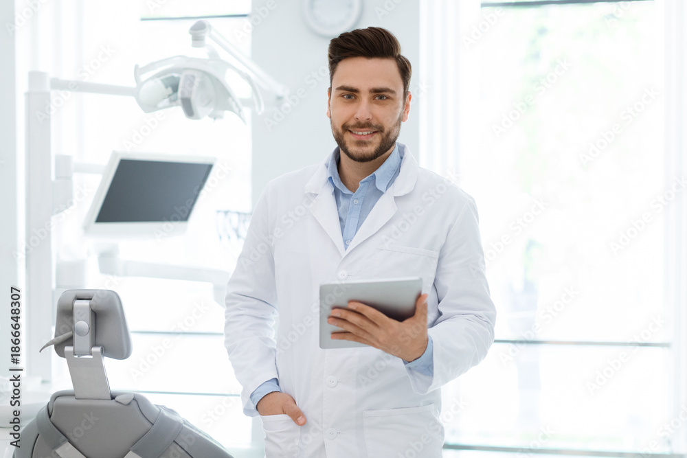 Fototapeta premium Attractive young bearded man dentist keeping beautiful smile on his face and holding digital tablet over cabinet background, empty space. Online application at dentist clinic concept