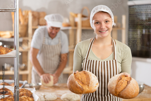 Billede på lærred Smiling young female baker holding golden loaves of crusty bread, proudly presenting freshly baked goods in cozy private bakery environment
