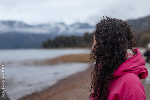 A female traveler with dark curly hair, wearing a pink hooded jacket, stands