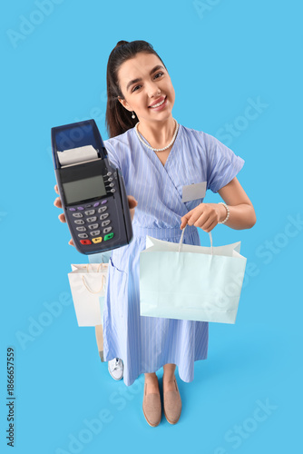 Young woman with shopping bag and payment terminal on blue background