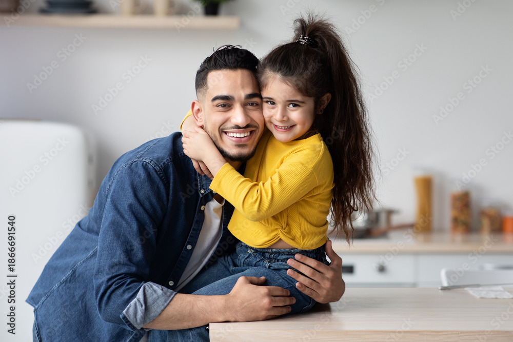 Obraz premium Portrait Of Happy Young Arab Dad And Little Daughter Posing Together In Kitchen Interior, Cute Small Girl Embracing Her Middle Eastern Father And Smiling At Camera, Daddy And Kid Having Fun At Home